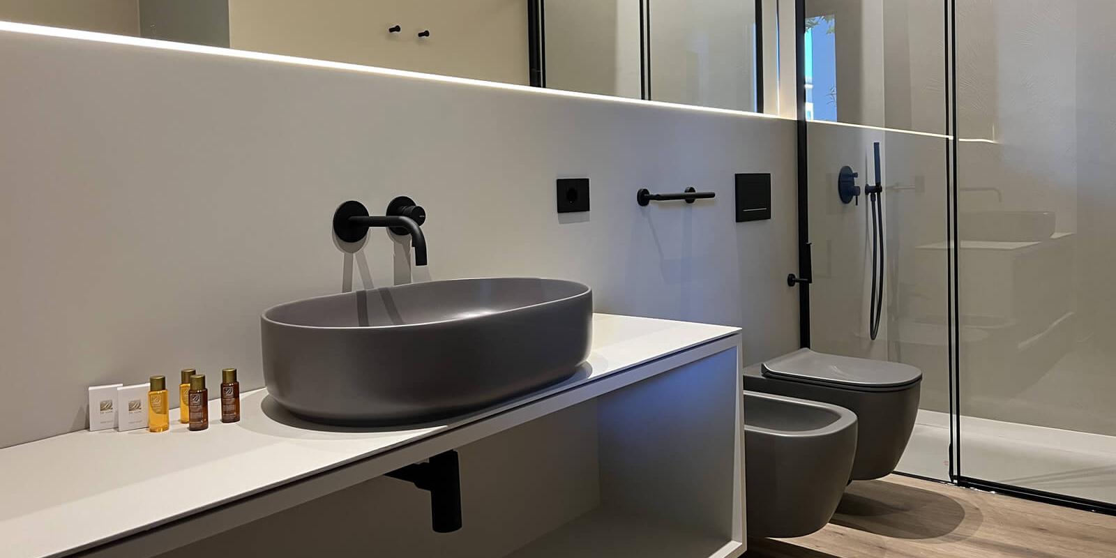 Bagno moderno con lavabo grigio, doccia in vetro e pavimento in legno chiaro.